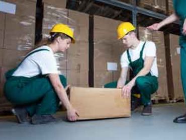 Two supervised workers lifting a cardboard box off the floor.